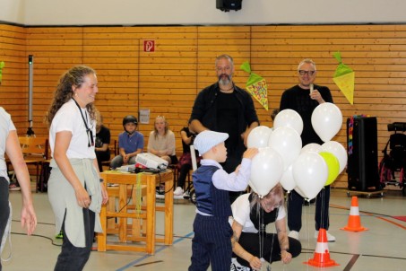 Junge von hinten zu sehen spielt mit weißen Luftballons. Zusätzlich sind zwei Frauen und ein Mann zu sehen