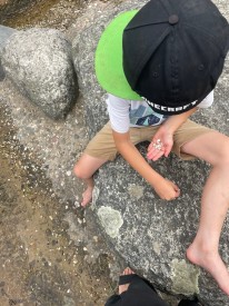 Junge mit schwarzer Capie ohne sichtbares Gesicht sammelt Steine am Bachlauf
