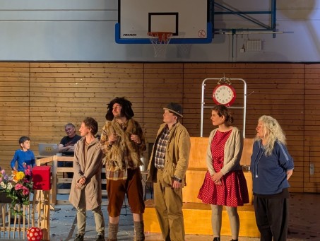 Theatergruppe steht nebeneinander. 3 Männer und 2 Frauen. Im Hintergrund sieht man einen Schüler und einen Mann