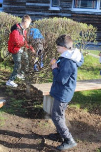 Ein Junge steht mit Osterei in der Hand vorm Geb&uuml;sch. 2 Kinder suchen im Geb&uuml;sch.